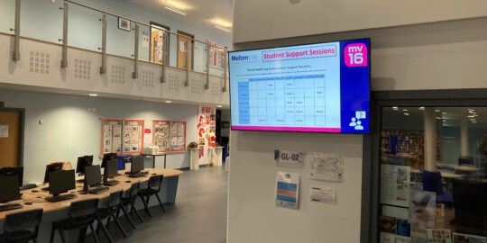 A school hallway with a digital display showing a school schedule hanging from the wall.