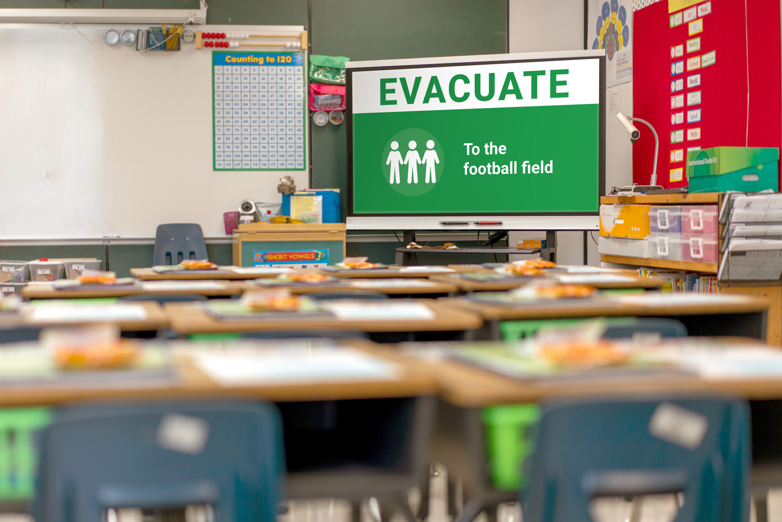 Classroom emergency evacuation alert displayed on a screen, instructing students to evacuate to the football field, with student desks and educational materials in the foreground.