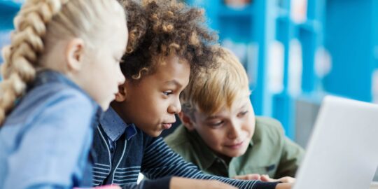 Group of three excited diverse students using laptop sitting at desk in school library. Mixed race schoolboy and his friends browsing through information on Internet