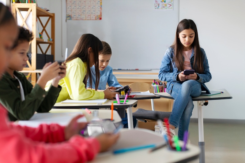 Elementary-age school children using cellphones while sitting at desks in a classroom.