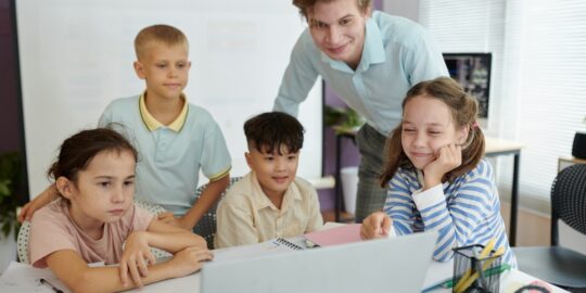 A group of elementary students looking at a laptop with a teacher looking on