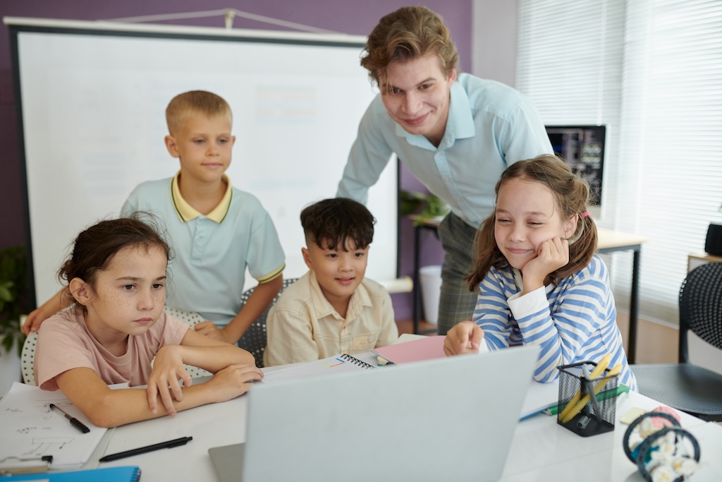 A group of elementary students looking at a laptop with a teacher looking on