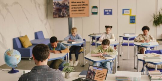 Students taking a test while a teacher looks on and mirrors his laptop screen to the classroom display.