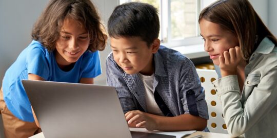 Diverse happy school kids using laptop computer together in classroom. Multicultural children junior students classmates learning online elementary education program class gathered at desk.