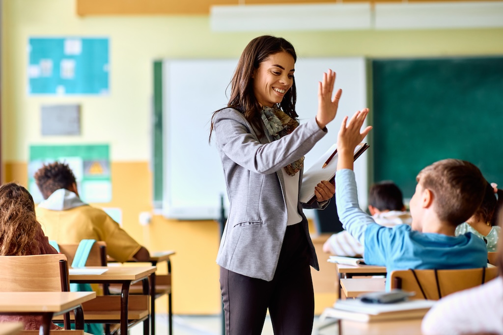 Happy elementary school teacher gives high-five to her student during class in classroom.